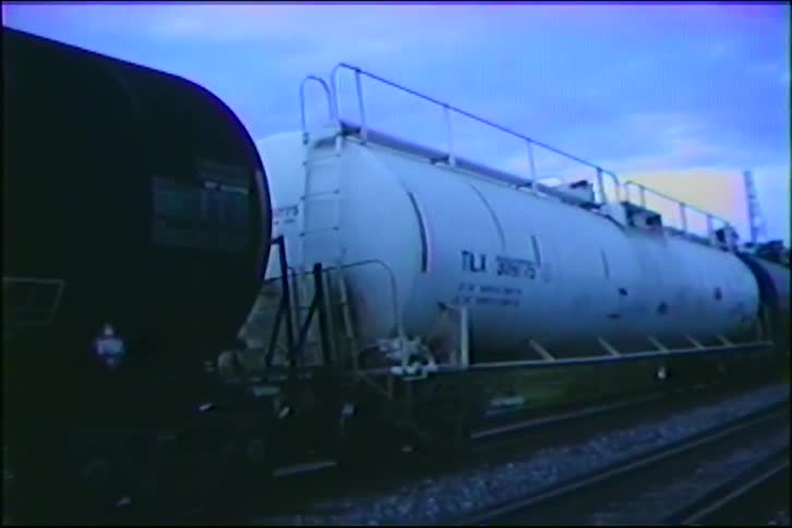 A freight train with various tanker cars moves slowly along the tracks at dusk, captured on vintage VHS tape