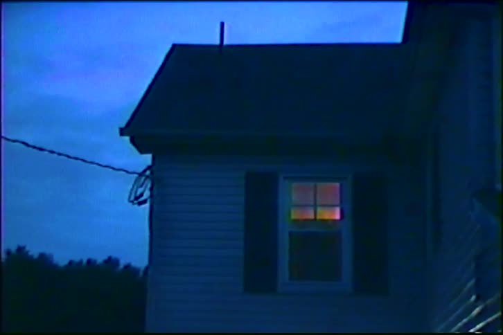 A vintage VHS shot shows a single illuminated window on the side of a house at dusk