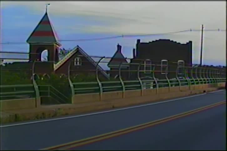 A vintage VHS style shot of a silver SUV driving over a bridge past a unique brick building at dusk