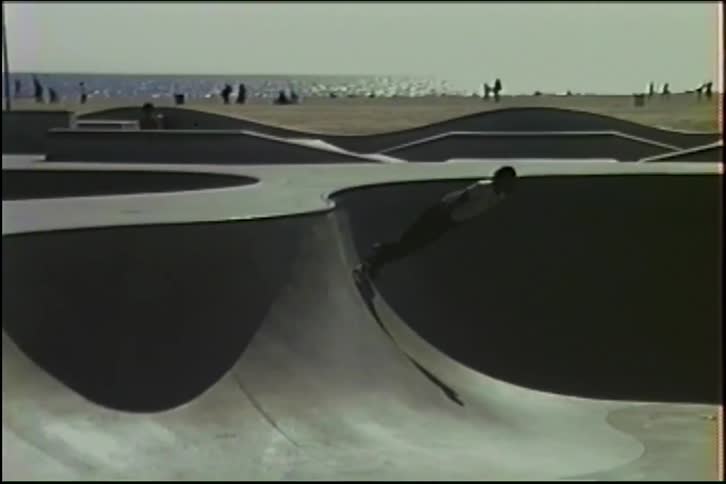 Vintage footage of a skateboarder carving through a concrete skatepark bowl next to the ocean on a sunny day