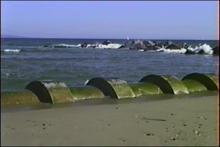 Slow motion waves crash against a rock jetty behind a moss-covered concrete drainage pipe on a sandy beach
