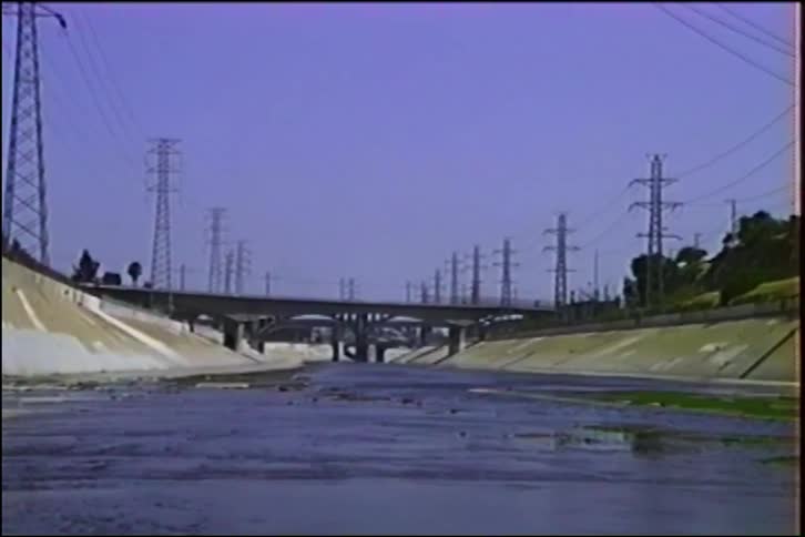 A wide shot from within the Los Angeles River channel shows a train crossing a concrete bridge, surrounded by power pylons under a pale sky