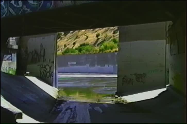 A static shot from a dark, graffiti-tagged concrete drainage channel reveals a slowly flowing river and sunny hillside beyond
