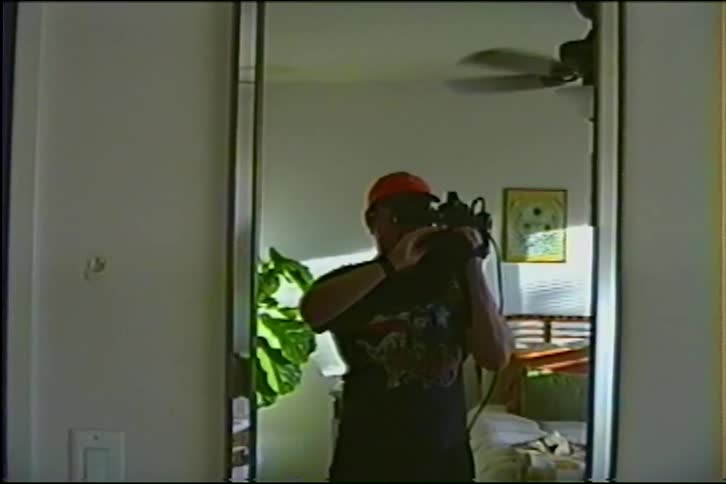 A man holding a vintage VHS camera films his reflection in a bedroom mirror and gives a peace sign