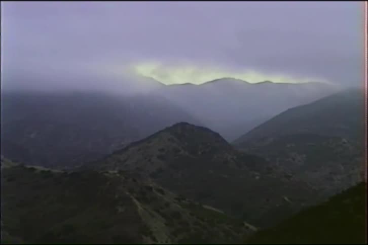 A slow zoom into a dark mountain range shrouded in thick fog, with a distant ridge illuminated by a break in the overcast sky