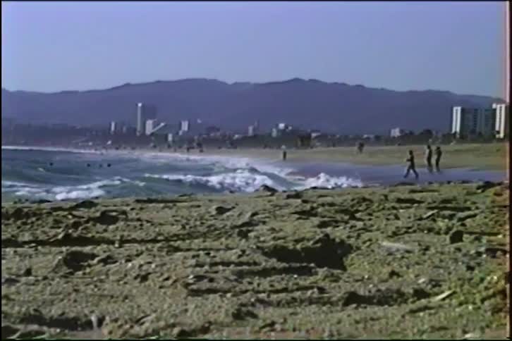 Vintage VHS footage zooms out from crashing waves to a wide view of a sunny city beach with distant mountains