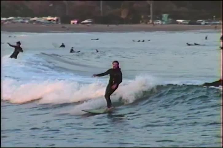 Vintage slow-motion footage shows a longboarder riding a wave among a crowd of other surfers