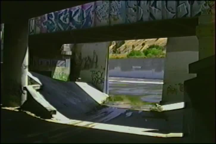 A slow pan reveals the graffiti-covered concrete structure of an urban underpass above a drainage ditch