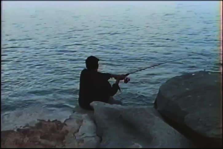 A lone fisherman sits peacefully on a rocky shoreline, casting and reeling in his line against the vast, calm water
