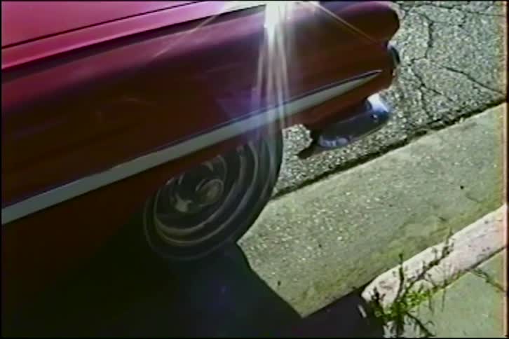 A bright sun flare reflects off the shiny red paint and chrome wheel of a vintage car parked on the street