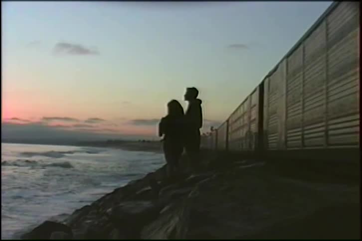 A silhouetted couple stands on coastal rocks watching the sunset as a graffiti-covered freight train rushes past