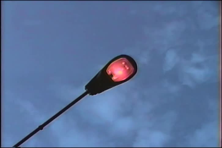 A silhouetted street lamp turns on and flickers with a red glow against a cloudy twilight sky