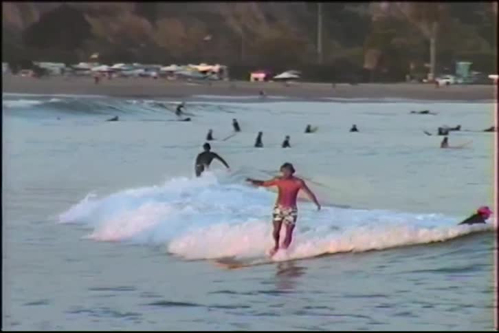 Vintage slow-motion footage of a man gracefully riding a longboard on a wave at a crowded beach, with a surprising appearance by a surfing dog