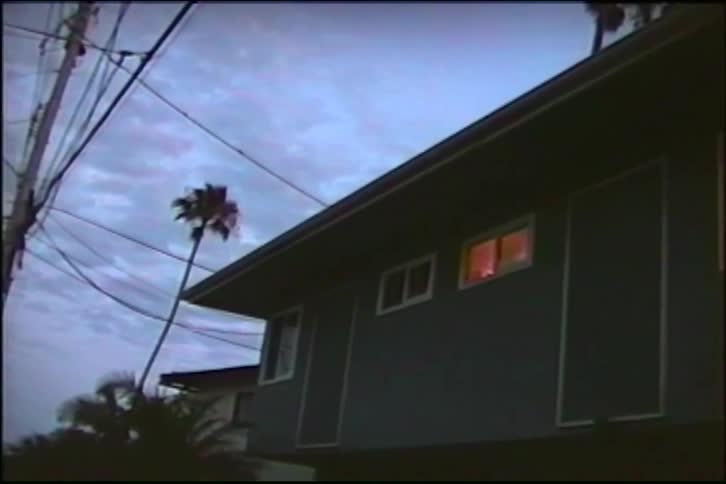 A static shot from a vintage home video shows a suburban house at dusk with a single glowing window and a palm tree against a cloudy sky