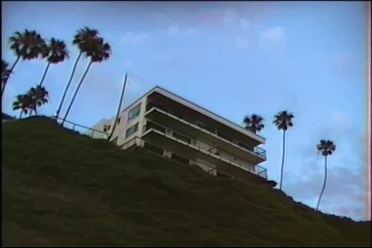 Vintage VHS footage of a modern house perched on a grassy hill surrounded by tall palm trees