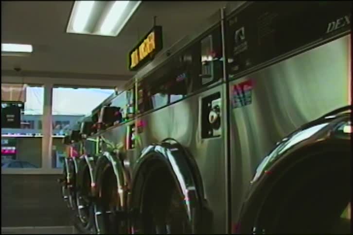A row of stainless steel washing machines spin clothes in a laundromat during the day