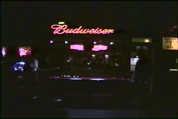 Two people play a game of pool in a dimly lit bar under a red neon Budweiser sign