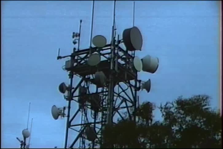 A static low-angle shot of a telecommunications tower covered in antennas and dishes against a bleak, overcast sky
