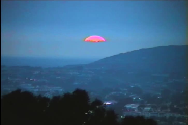A vintage VHS shot shows a vibrant pink sun setting over a hazy coastal town nestled in rolling hills at dusk