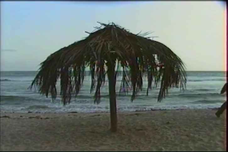 A long-haired man playfully runs circles around a tiki-style umbrella on the beach with the ocean in the background