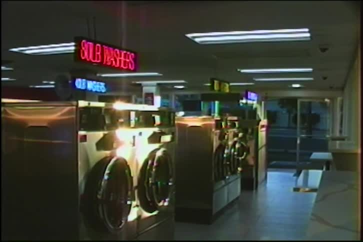 VHS footage captures the quiet, empty interior of a retro laundromat, zooming in on the glowing red and blue neon signs