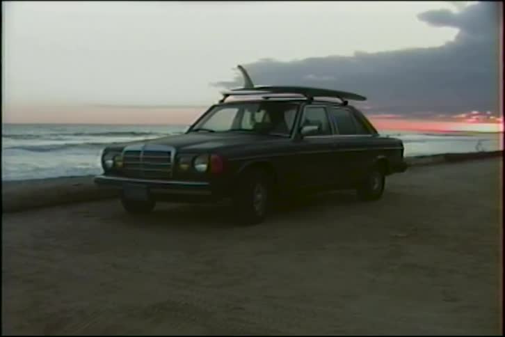 A vintage Mercedes-Benz with a surfboard strapped to the roof is parked on a sandy beach at sunset