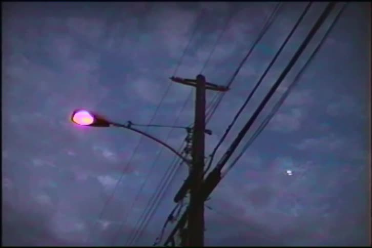 Lo-fi footage of a pink streetlight flickering to life on a utility pole against a moody, cloudy evening sky