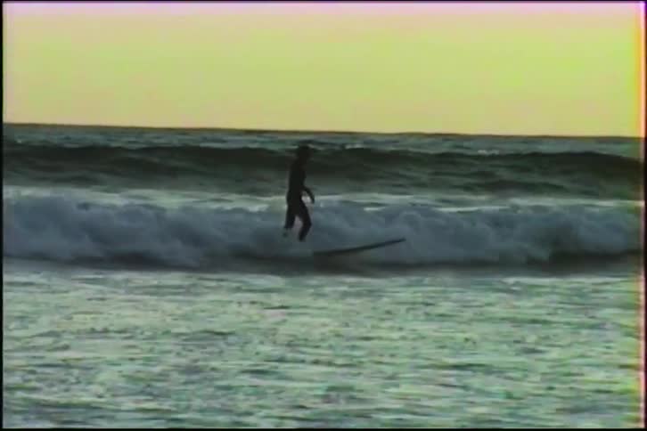 A silhouetted surfer skillfully rides a wave in slow motion against the twilight sky in this vintage footage