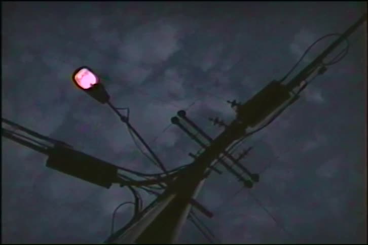 A street light on a silhouetted telephone pole turns on against a moody, dark sky at dusk