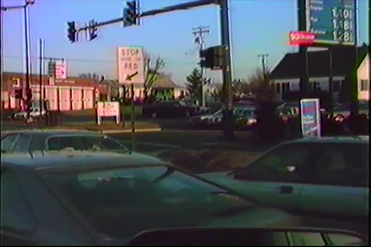 A grainy, vintage home video captures a view of an Exxon gas station and traffic at an intersection, filmed from inside a car in the 1980s