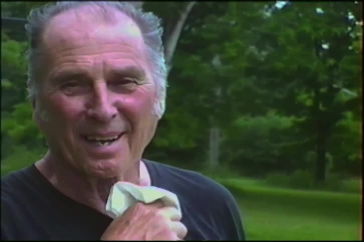 An elderly man waves and smiles warmly at the camera while standing in his backyard in this vintage home video