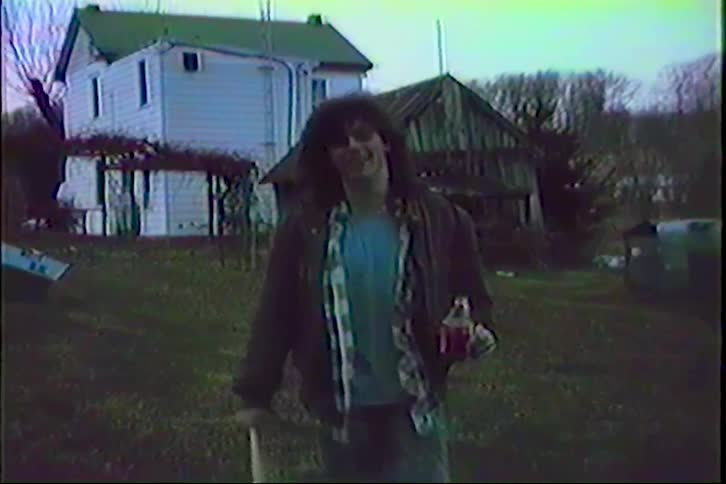 Vintage VHS footage shows a long-haired young man in 80s attire walking towards the camera in a rural backyard while holding a soda bottle