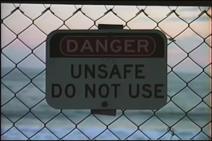 A vintage VHS-style shot zooms in on a 'Danger Unsafe Do Not Use' sign attached to a fence with ocean waves in the background at sunset