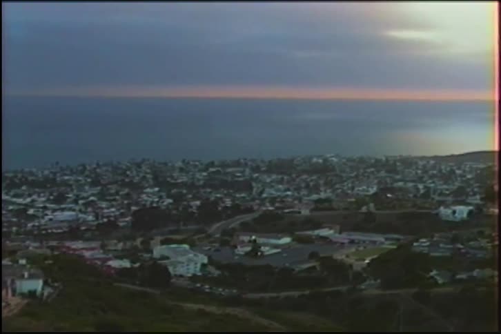 A slow pan across a California coastal city from a hillside vantage point during a hazy sunset, filmed on vintage tape