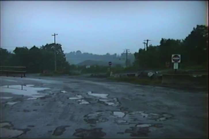 A desolate, wet rural road filled with potholes reflects the gloomy sky on an overcast day