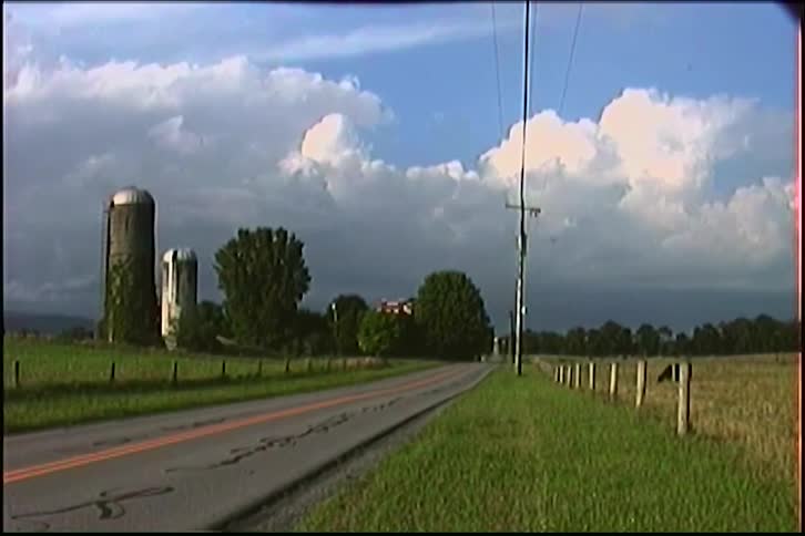 A static shot from a vintage camera of a rural country road leading past a farm with silos under a dramatic, cloudy sky