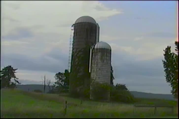 Old, vine-covered farm silos stand on a rural hillside under a cloudy sky in this nostalgic, vintage footage