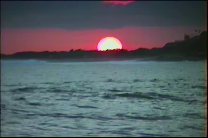 A vintage shot of a vibrant pink and red sunset over the ocean horizon as waves gently roll in