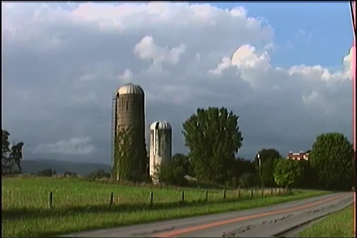 Two weathered farm silos stand in a green field next to a country road under a dramatic sky