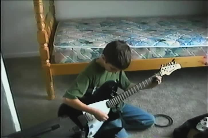 A young boy sits on his bedroom floor and practices playing his electric guitar in a grainy home video from the 1990s