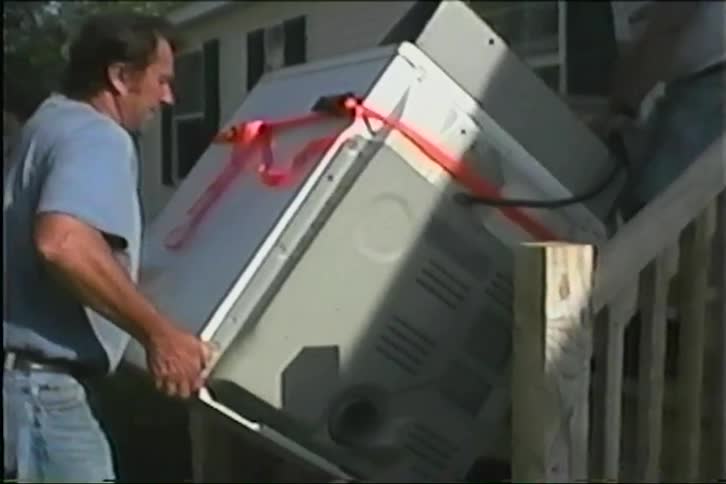 Two men use a dolly to carefully move a large clothes dryer down a set of outdoor wooden stairs