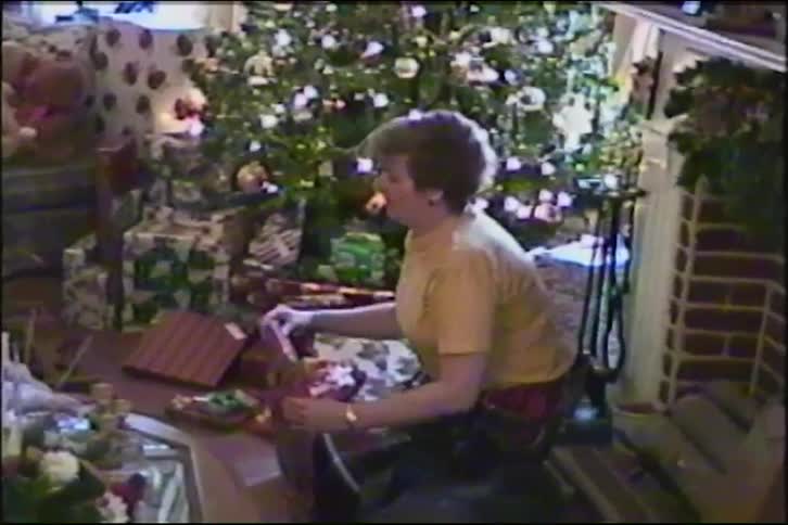 A woman wraps Christmas presents on the floor of her living room in front of a beautifully decorated tree and a cozy fireplace