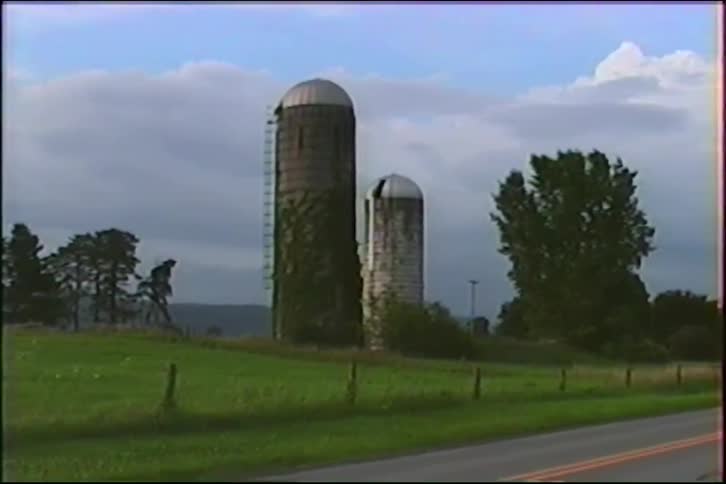 A vintage VHS shot captures two weathered farm silos, one covered in ivy, standing on a green hill next to a country road
