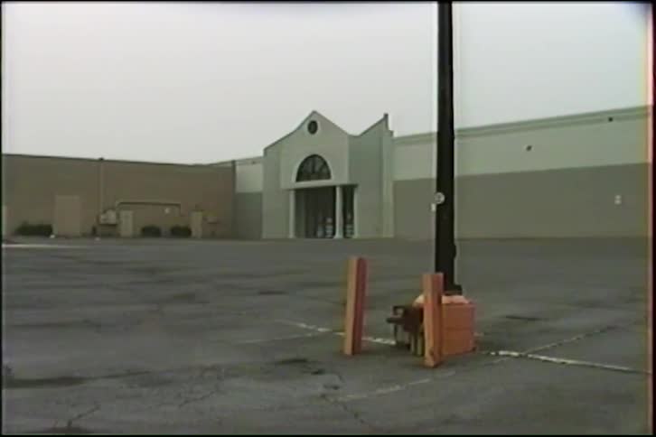 Vintage VHS footage shows a desolate, empty parking lot in front of a large retail store on an overcast day
