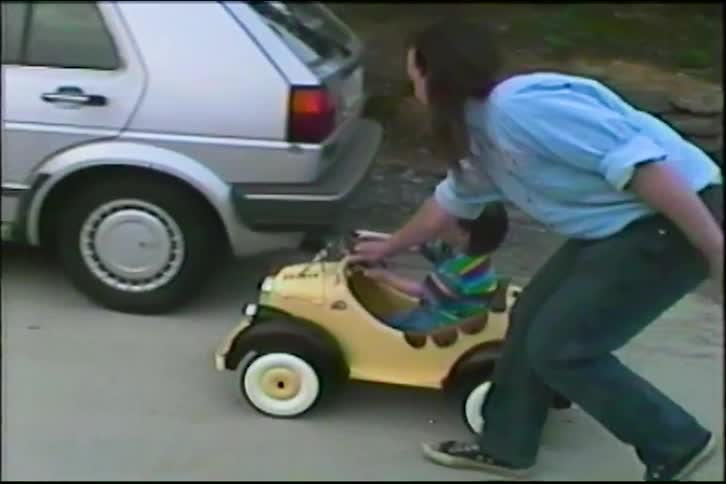 A father with a mullet helps his young son drive a vintage-style yellow toy car on a suburban driveway