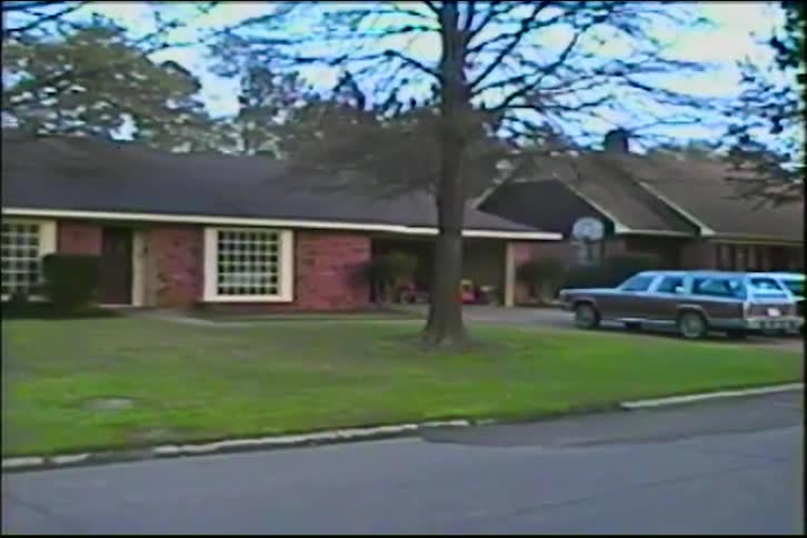 A vintage home video captures a slow pan of a suburban brick house with a station wagon in the driveway