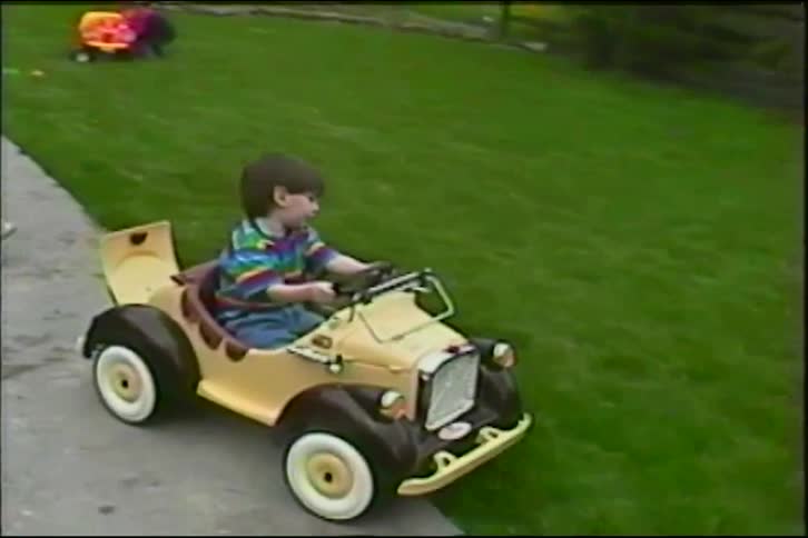 Vintage home movie shows a father helping his young son ride in a classic toy car down a sidewalk in their yard