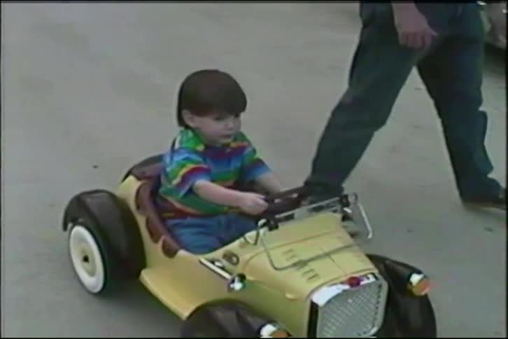 A father walks alongside his young son who is driving a vintage yellow toy car in this charming home video