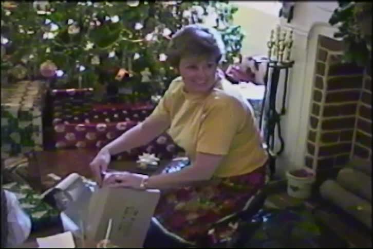 A happy woman laughs while wrapping a Christmas present in front of a decorated tree in a vintage home video