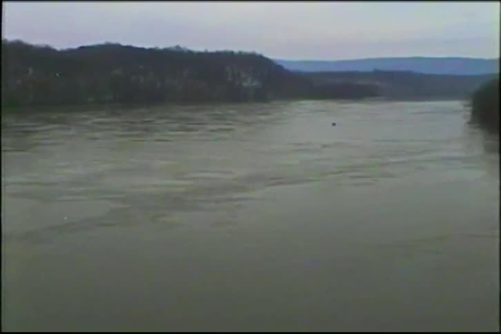 A wide, murky river flows past a forested, hilly shoreline on a cold, overcast day